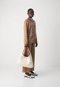 Braun gemusteter Anzug mit Knöpfen, kombiniert mit einer großen weißen Handtasche. Das Modell trägt schwarz-weiße Schuhe mit dekorativen Akzenten.
