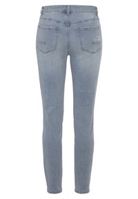 Jean en denim bleu clair, coupe ajustée, taille mi-haute, poches arrière avec patch, et un détail de logo discret au-dessus de la poche droite.