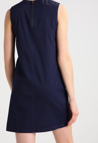 Světlogradá modrá bezramínková šaty s vysokým výstřihem, zipem na zadní části a rovným střihem padá nad kolena.