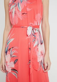 Robe corail avec un motif floral, en tissu léger, et une ceinture nouée à la taille. Affiche des fleurs roses vives avec des accents verts.