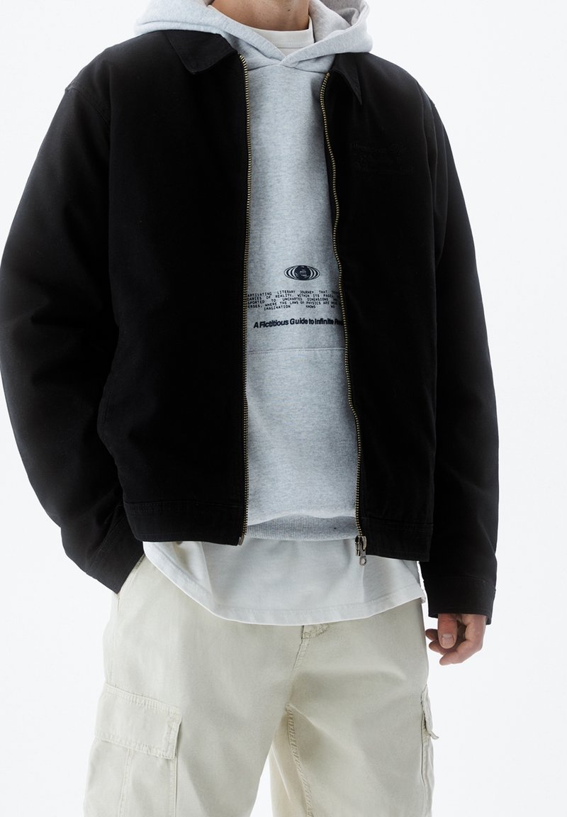 Schwarze Zip-Jacke mit Kragen, über einem grauen Hoodie mit Text getragen, kombiniert mit hell-beigen Cargo-Hosen. Einfaches, lässiges Design.