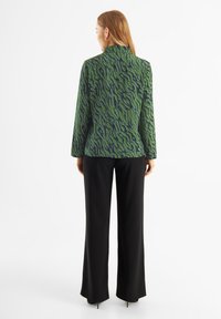 Giacca a maniche lunghe di colore verde con un motivo ondulato, abbinata a pantaloni neri a gamba larga. Tessuto morbido, design senza colletto, silhouette aderente.