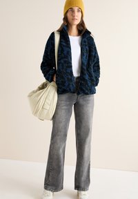 Giacca zip-up morbida blu navy e nera, camicia bianca, jeans grigio chiaro e un cappellino di lana giallo. Tiene una borsa beige. Sneakers bianche.