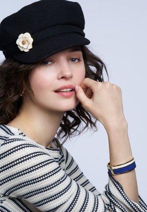 Jeune femme portant un chapeau noir avec une épingle à fleur blanche, un pull rayé et un bracelet bleu et blanc, posant avec sa main près de ses lèvres.