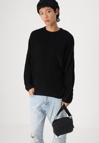 Suéter negro de canalé con hombros caídos, combinado con jeans desgastados de color azul claro. En una mano, sostiene un pequeño bolso negro con asa.