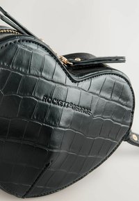Borsa a forma di cuore nera realizzata in finta pelle di coccodrillo testurizzata. Caratterizzata da accessori in metallo dorato e chiusura con zip. Nome del marchio in rilievo.
