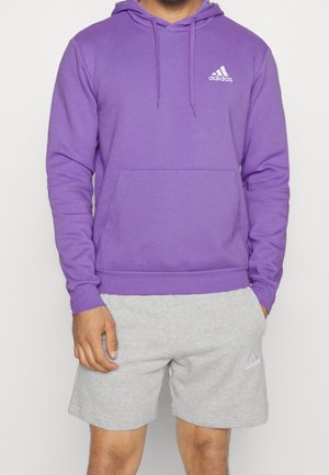 Muž v fialové mikině Adidas a světle šedých šortkách Adidas stojí s jednou rukou v kapse šortek před jednobarevným pozadím.