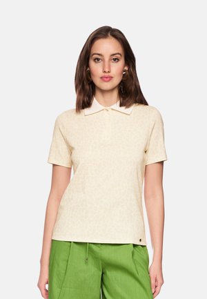 Femme aux cheveux bruns mi-longs portant un polo beige à manches courtes et un pantalon vert, debout devant un fond uni.