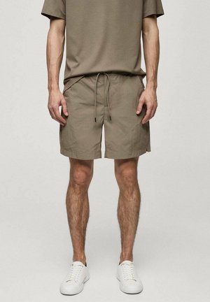 Hellbraune Shorts mit elastischem Bund und Kordelzug. Hergestellt aus weichem Stoff, mit seitlichen Taschen und einem lässigen Schnitt. Getragen mit weißen Sneakers.