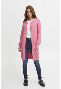 Roze cardigan met een lange, open voorkant, geribbelde textuur en lange mouwen. Gecombineerd met een witte top en donkerblauwe skinny jeans.