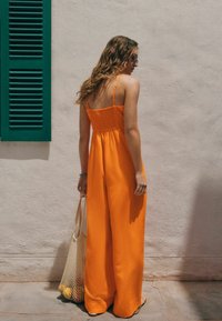 Salopette orange avec un dos smocké, des bretelles ajustables et un design à jambes larges. Un sac en filet pend d'une main, avec un mur texturé en arrière-plan.