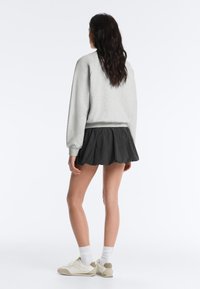 Femme debout, dos tourné, portant un sweat-shirt gris clair, une mini-jupe plissée noire, des chaussettes blanches et des baskets beiges sur fond blanc.