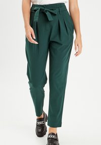 Femme portant un pantalon taille haute vert foncé avec une ceinture en tissu nouée et des mocassins noirs avec des chaînes en argent, se tenant devant un fond uni.