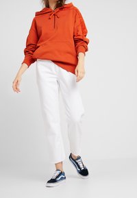Oranje pullover-hoodie met trekkoord, ritssluiting bij de nek en voorvak; gecombineerd met witte cropped jeans en marineblauwe sneakers met blauwe accenten.