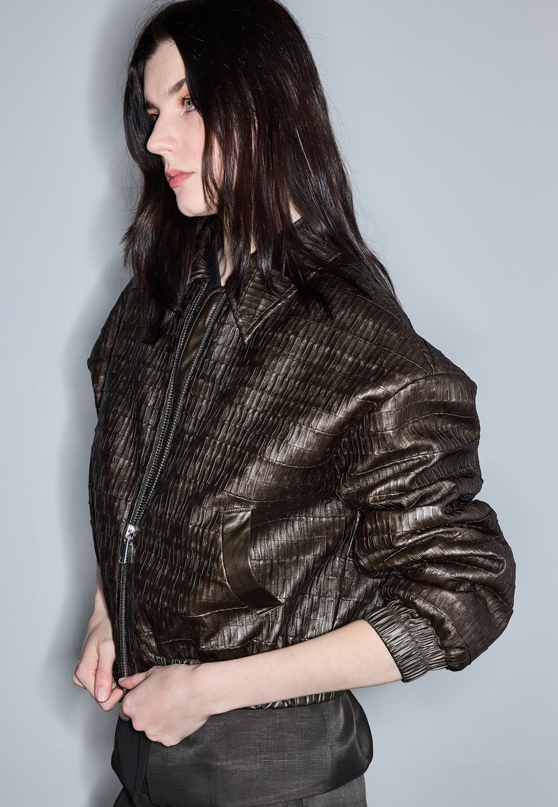 Woman with dark hair wears a textured dark brown leather jacket, standing sideways against a plain light gray background.