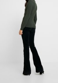 Pull en maille côtelée vert foncé avec une silhouette ajustée, associé à un pantalon noir évasé en tissu texturé, vue de dos.