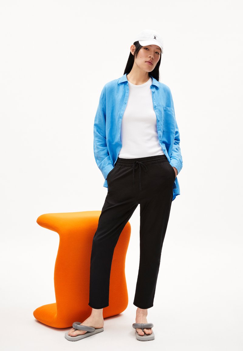 Junge Frau trägt ein blaues Hemd, eine weiße Kappe, ein weißes Oberteil, schwarze Hosen und graue Sandalen und steht neben einem leuchtend orangefarbenen modernen Stuhl.