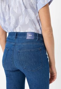 Jeans in denim blu medio con una texture liscia, dotati di una tasca posteriore quadrata e di un'etichetta blu. Indossati con un top chiaro a fantasia.
