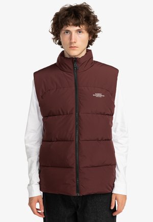Weinrote Steppweste mit hohem Kragen, Reißverschluss vorne und horizontalen Nähten. Getragen über einem weißen Langarmshirt mit dunklen Hosen.