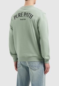 Groene sweatshirt met ribgebreide manchetten en onderkant, met zwarte opdruk "PURE PATH Social Club" op de achterkant. Draag het met lichtblauwe jeans.