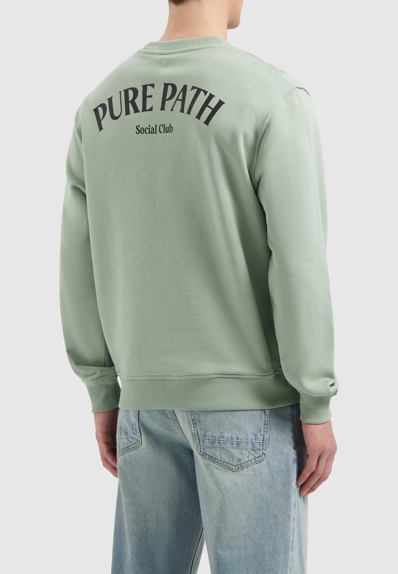 Groene sweatshirt met ribgebreide manchetten en onderkant, met zwarte opdruk "PURE PATH Social Club" op de achterkant. Draag het met lichtblauwe jeans.
