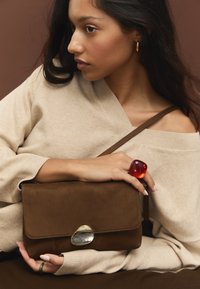 Rjava torbica iz semša z zaponko iz kovine, ki jo drži oseba v bež puloverju, na eni roki pa ima velik rdeč prstan.