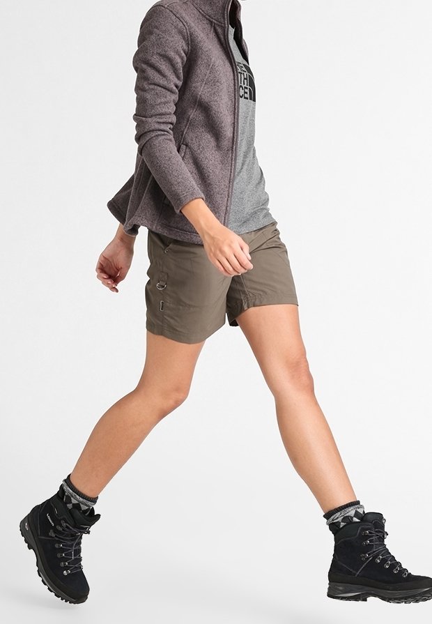 Persona caminando con botas de senderismo, pantalones cortos caqui, camiseta gris y una chaqueta morada con cremallera contra un fondo blanco.