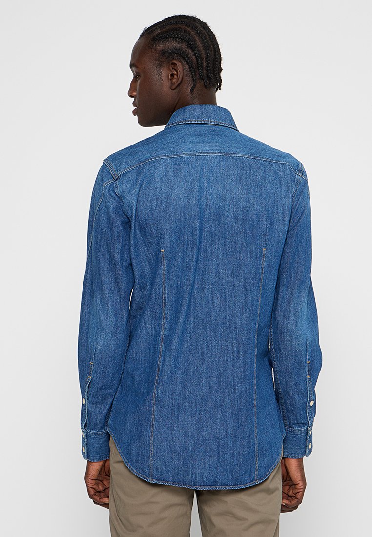 Homme avec une coiffure en nattes africaines portant une chemise en denim bleu à manches longues et un pantalon beige, tourné de dos devant un fond uni.