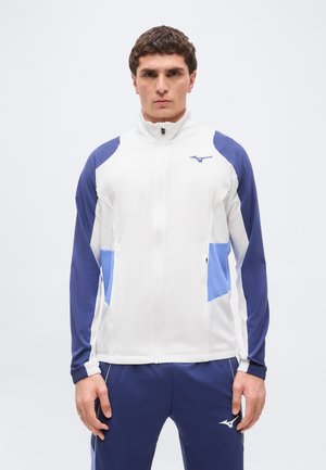 Man standing facing forward wearing white and blue zip-up sports jacket and navy blue athletic pants with logos on chest and thigh.