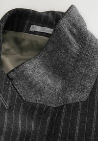 Grijze wollen revers met een textuur afwerking, voorzien van een donker streepjespatroon. Binnenvoering van glad, lichtgekleurd stof.