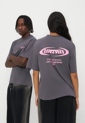 Deux modèles portent des t-shirts gris oversize avec un texte rose "URBAN Your Turn Studio Urban Streetwear", l'un de face, l'autre montrant le dos.