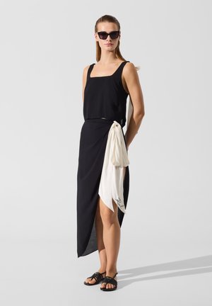 Kvinde iført sort ærmeløs top og asymmetrisk sort nederdel med hvidt stofdetalje, sorte sandaler og mørke solbriller, stående.