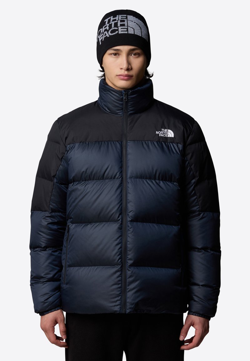 Mörkblå puffjacka med svarta accenter, hög krage, dragkedjestängning och logodetalj på bröstet. Har quiltat mönster och elastiska ärmslut.