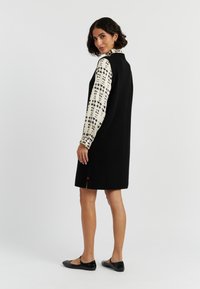 Schwarzes Wollkleid ohne Ärmel mit hohem Kragen, das am Saum einen roten Akzent hat und über einer weißen Bluse mit schwarzen Floralprint-Ärmeln getragen wird.