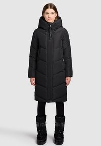 Svart lång vadderad jacka med hög krage, synliga sömmar och blixtlåsförsedda stängning. Bärs med svarta stövlar med snörningsdetaljer.