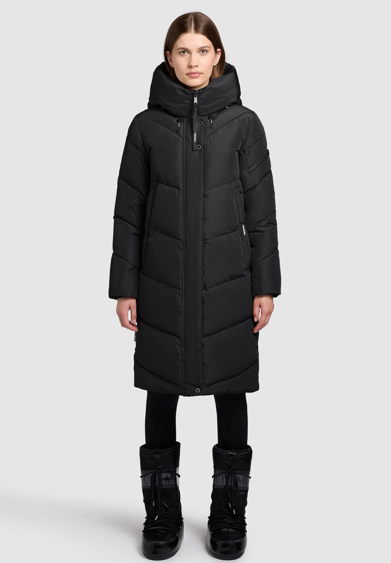 Czarna długa kurtka puchowa z wysokim kołnierzem, widocznymi przeszyciami i zamkiem błyskawicznym. Noszona z czarnymi butami sznurowanymi.
