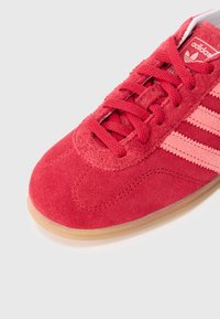 Sarkanu zamšādas sporta apavu ar teksturētu augšdaļu, kurā ir trīs gaišāk sarkanas svītras un gumijas ārējā zole. Šņores atbilst apavu krāsai.