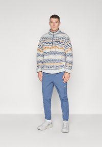 Πουλόβερ από φλις σε κρέμ χρώμα με μπλε και πορτοκαλί σχέδια, μισό φερμουάρ στον λαιμό. Συνδυασμένο με ανοιχτό μπλε παντελόνι και γκρι παπούτσια.