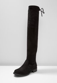 Bota de ante negra por encima de la rodilla con punta redondeada, diseño ajustado y un tacón bajo. Presenta un lazo ajustable en la parte superior.