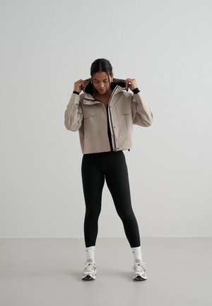 Naine reguleerib beežikat kapuutsiga jopet, kannab musti retuuse, valgeid sokke ja tossusid, seisab tavalise halli seina ning põranda taustal.