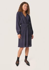 Navyblaues, langärmeliges Kleid mit elastischem Bund und Bindedetail, das einen tiefen V-Ausschnitt und einen glatten, leichten Stoff aufweist.