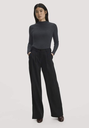 Vrouw staat met handen in de zakken, draagt een donker geribd top met lange mouwen, brede zwarte broek en donkere schoenen op een effen achtergrond.
