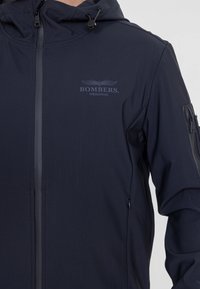 Navyblauwe hooded jack gemaakt van gladde stof, met een voorrits, logo op de borst en een ritszak op de mouw.