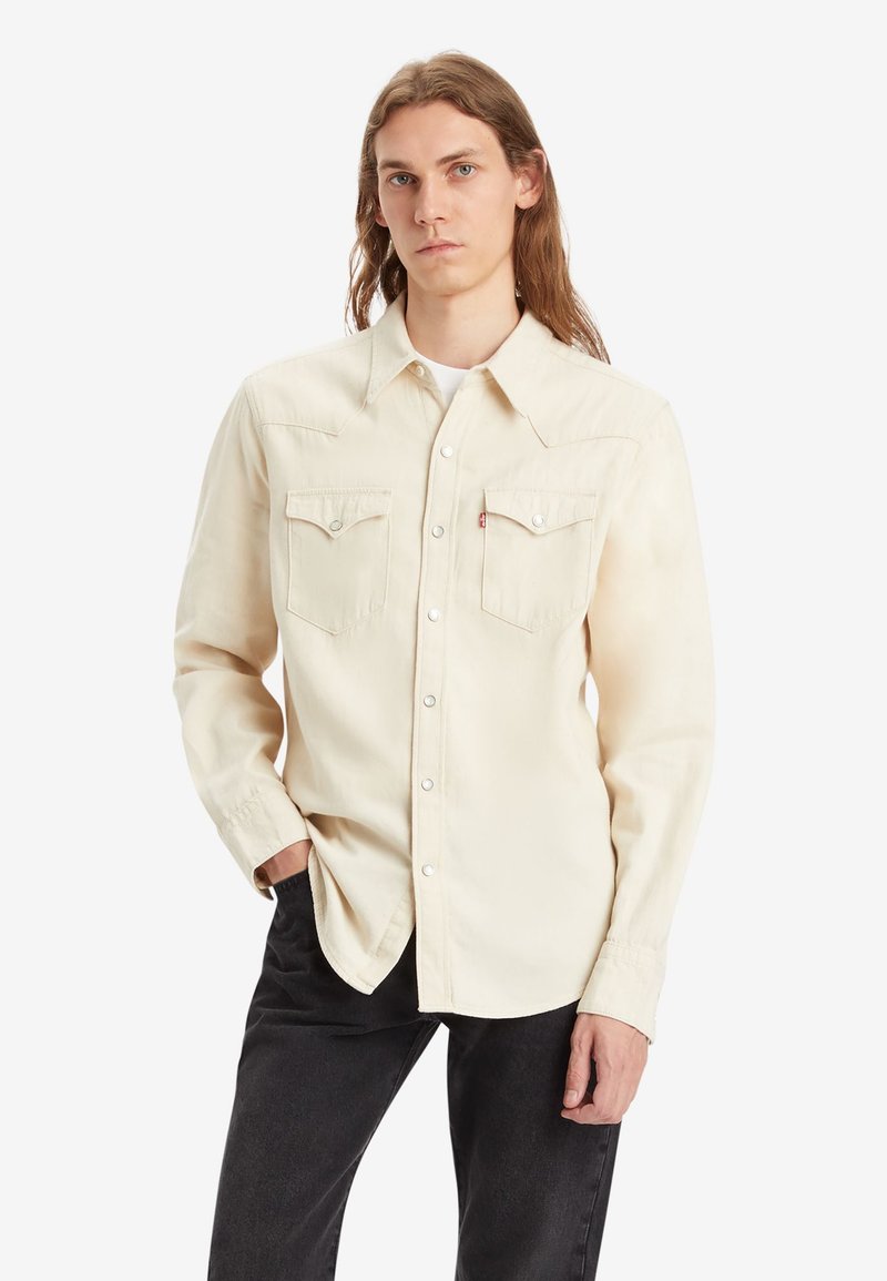 Camisa beige claro de botones con dos bolsillos en el pecho, botones a presión y detalles de yugo en estilo occidental. Manga larga con puños abotonados.