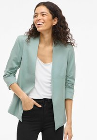 Femme souriante avec des cheveux bouclés, portant un blazer vert clair, un haut blanc et un pantalon noir, avec une main dans sa poche, sur un fond blanc.