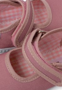 Las zapatillas de lona rosas presentan una forma redondeada, una tira elástica y un interior de tela a cuadros. Los acentos cosidos realzan el detalle estructural.