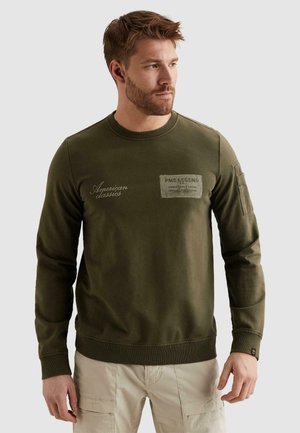 Moški z brado, oblečen v olivno zelen pulover s kapuco z napisom "American classics" in bež cargo hlače, gleda v desno na enobarvnem ozadju.