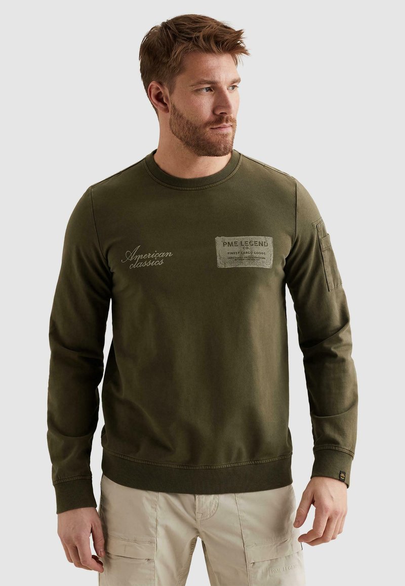 Homme barbu portant un sweat-shirt vert olive avec le texte "American classics" et un pantalon cargo beige, regardant vers la droite sur un fond uni.