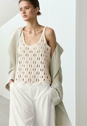 Junge Frau trägt ein beiges gehäkeltes Tanktop, lockere weiße Hose und eine leichte Jacke, die über die Schultern drapiert ist, drinnen.