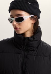 Giacca imbottita nera con collo alto, indossata con occhiali da sole trasparenti e angolari con lenti scure e dettagli pastello. Berretto nero lavorato a maglia sulla testa.